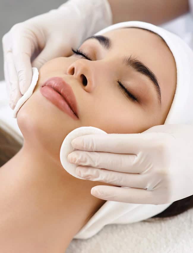 A woman with closed eyes receives a gentle cleansing treatment with cotton pads from a professional wearing white gloves, illustrating the relaxing and rejuvenating steps of a holistic facial.